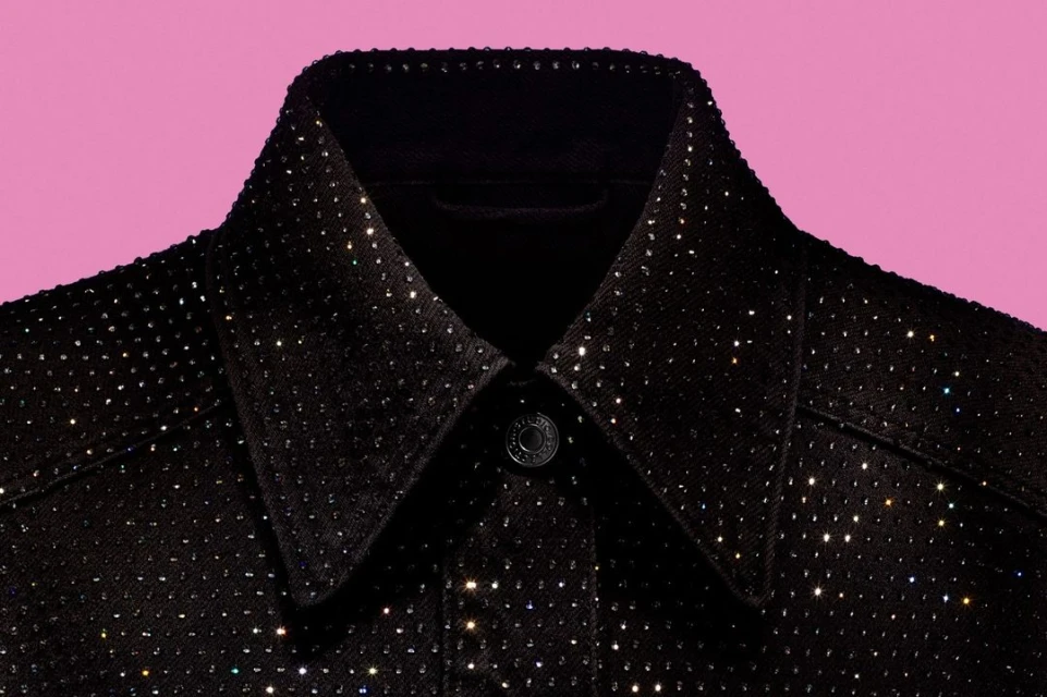 Close-up of a black denim jacket collar, adorned with sparkling rhinestones, set against a light pink background for fashion appeal.
