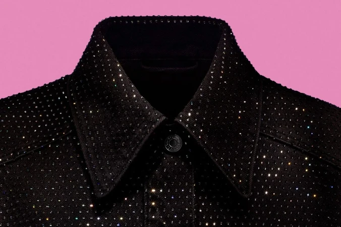 Close-up of a black denim jacket collar, adorned with sparkling rhinestones, set against a light pink background for fashion appeal.