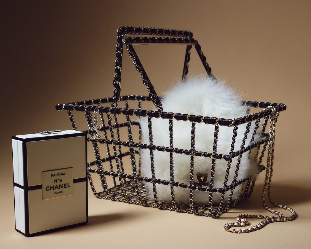 Chanel N°5 perfume box and metallic chain basket with white fur, presented against a tan background in a studio fashion shot.