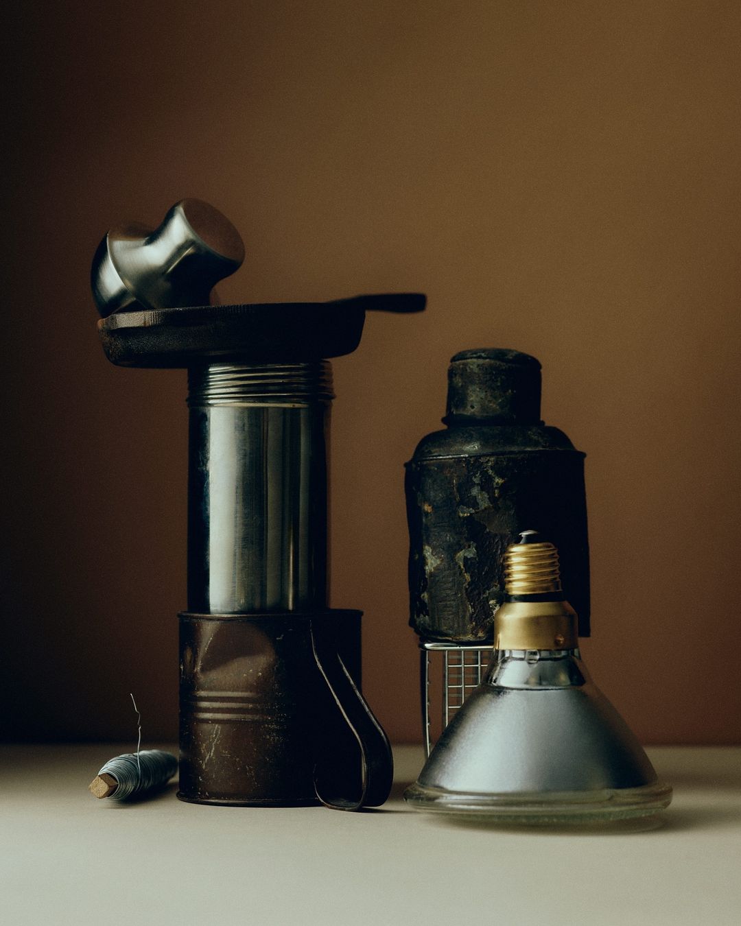 Still life composition featuring a stack of metal kitchenware including a frying pan, cans, saucepan, and lamp against brown background.