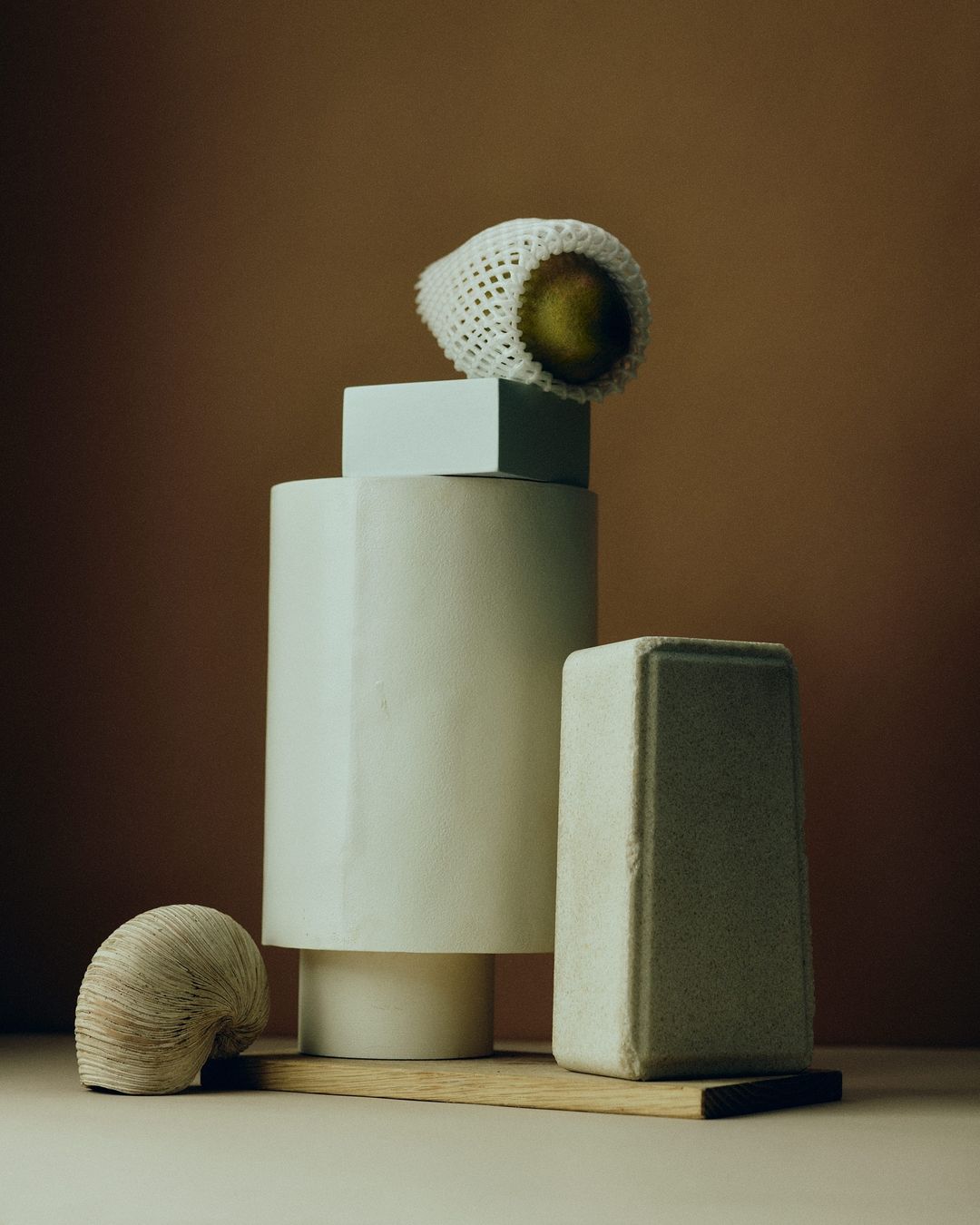 Still life featuring a pear in a net bag on stacked geometric shapes, with a neutral color palette and clean studio lighting.