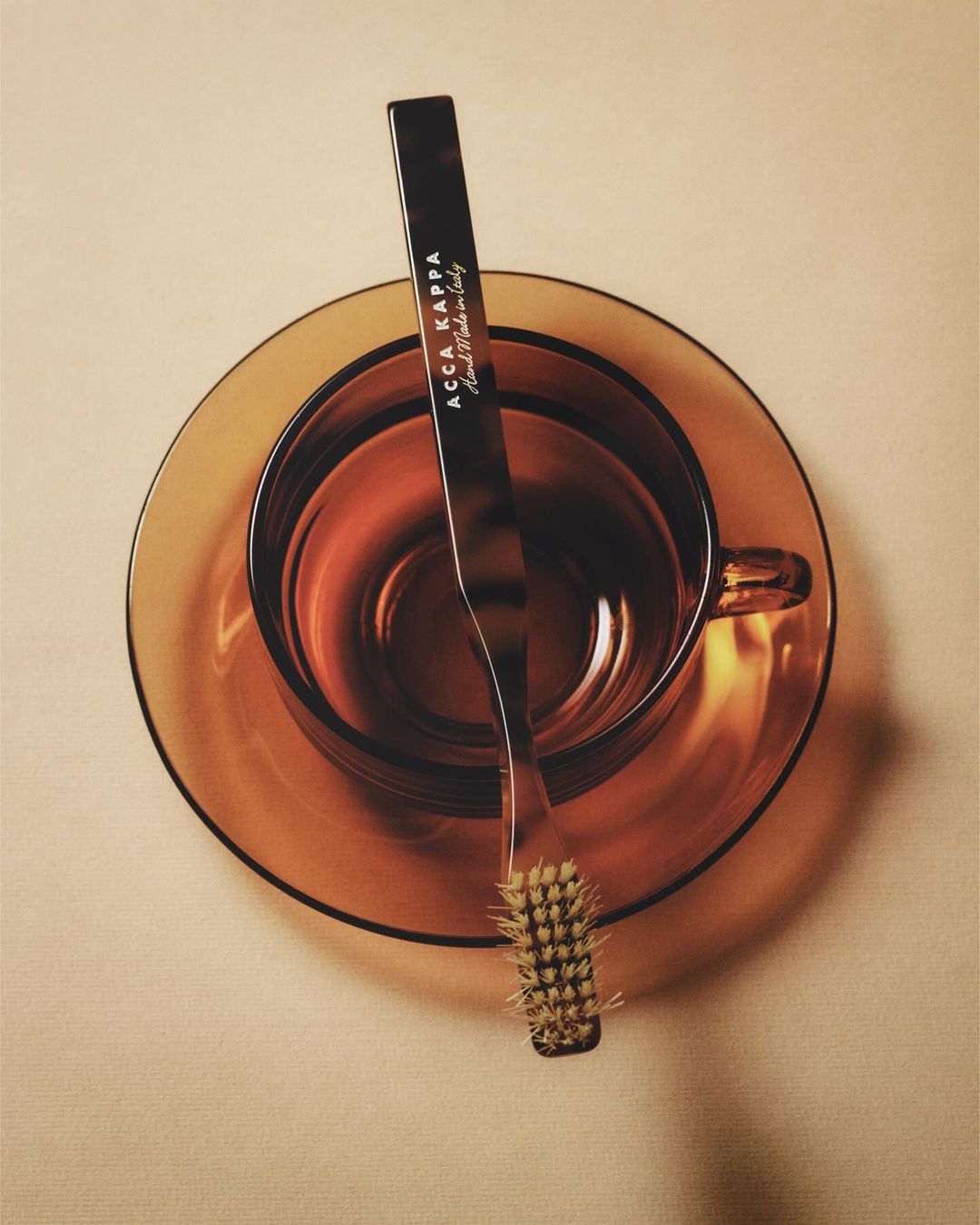Still life studio shot of an amber-colored glass cup and saucer with a small brush from Acca Kappa laying across the rim.