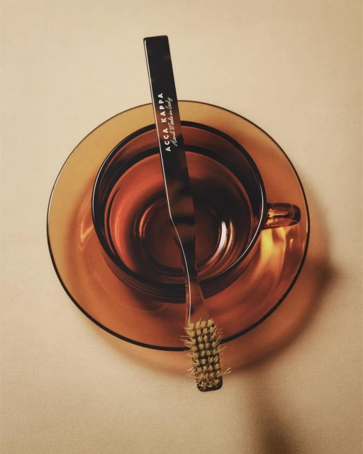 Still life studio shot of an amber-colored glass cup and saucer with a small brush from Acca Kappa laying across the rim.