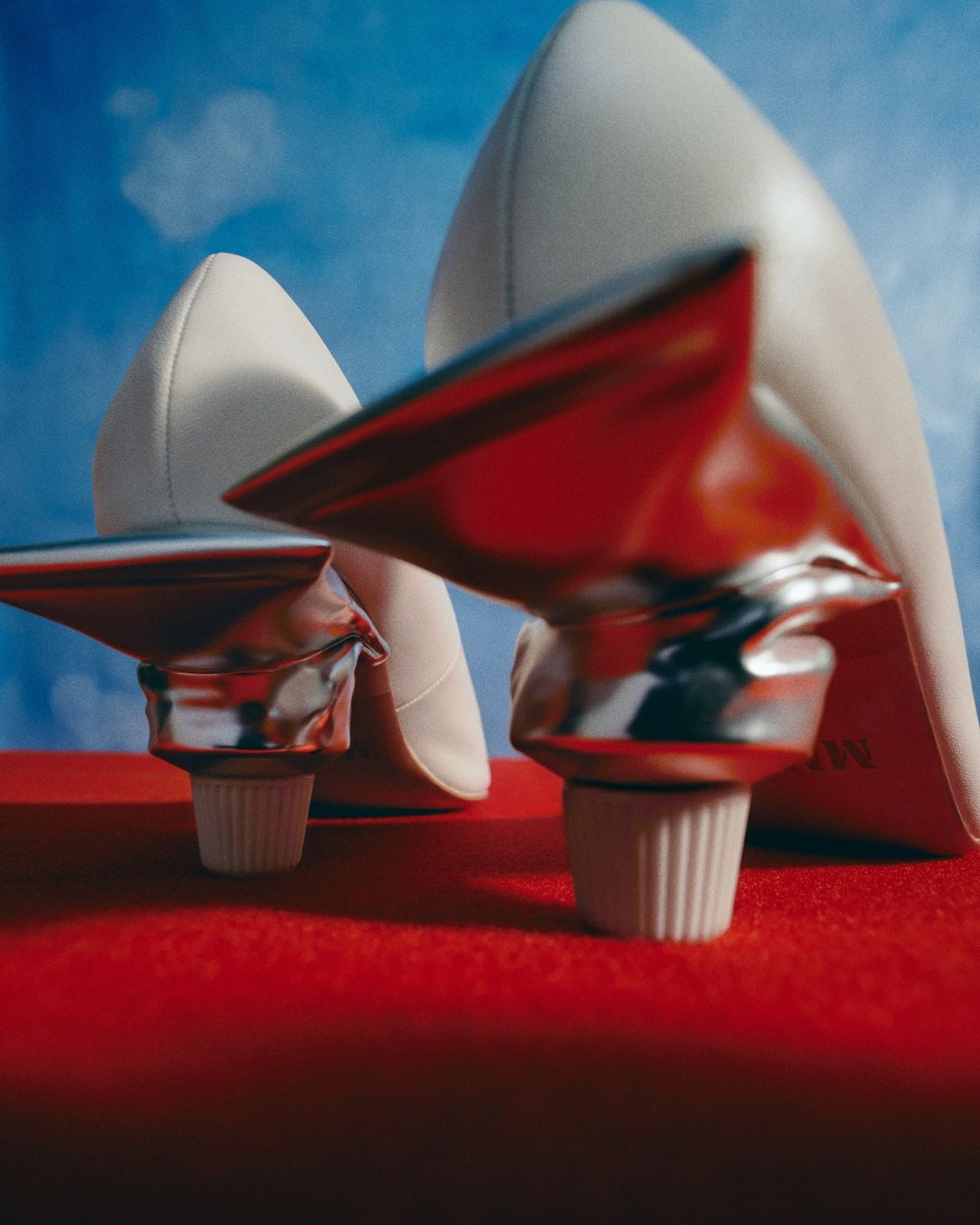 Elevated view of two white high heel shoes balanced on silver and white cosmetic tubes against a bright blue sky backdrop.