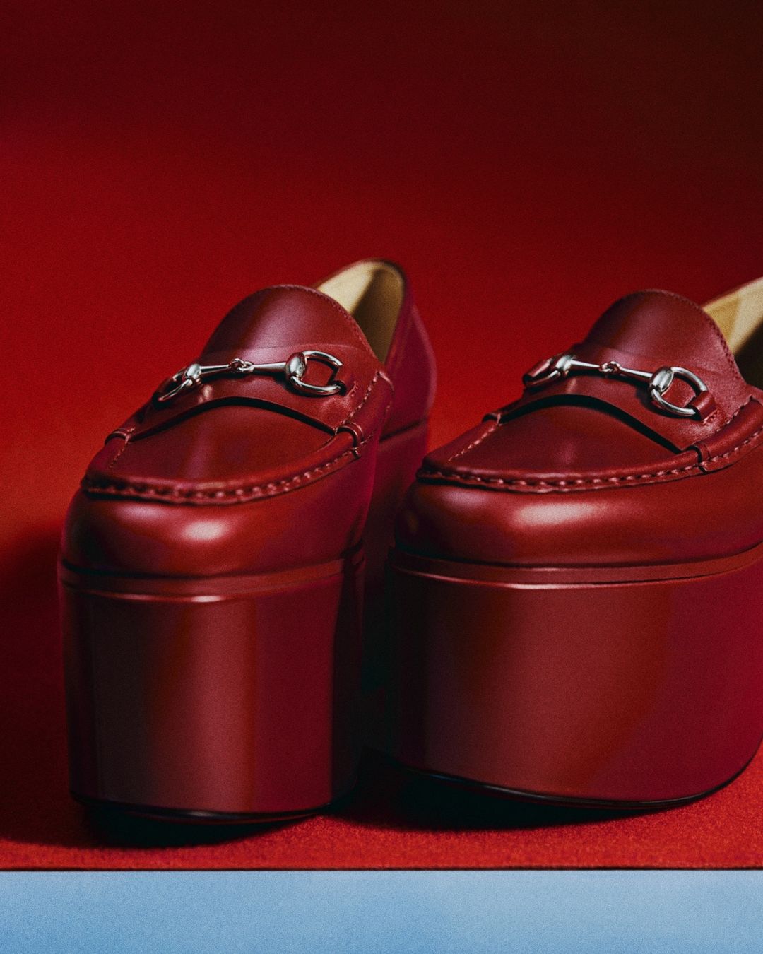 Close-up of a pair of platform loafer shoes in red leather with metal hardware, set against a backdrop of red and blue.