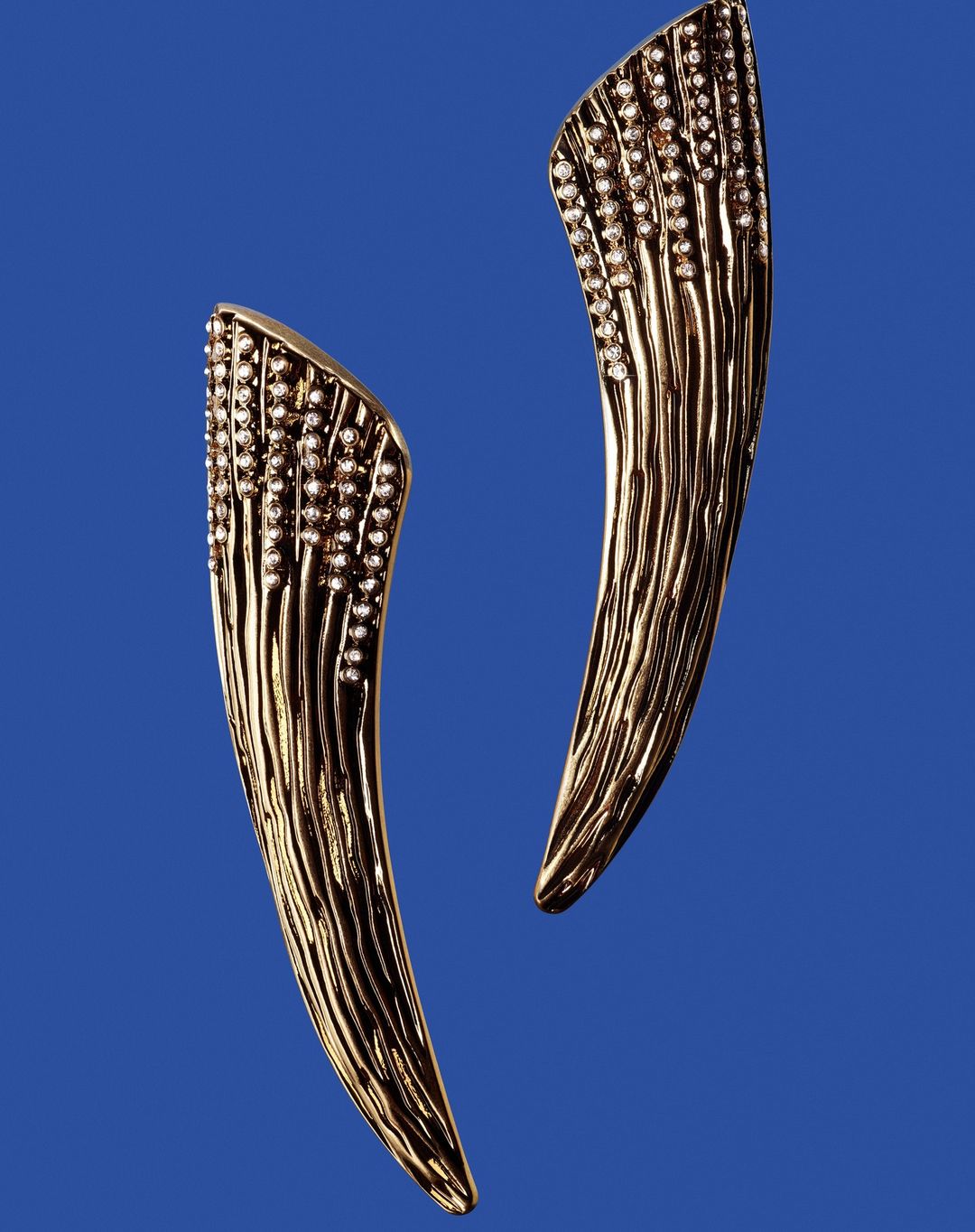 Two curved gold earrings encrusted with small diamonds displayed against a solid blue background.