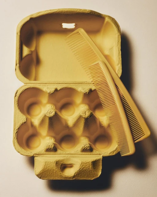 Close-up shot of two yellow combs resting on an open, empty yellow egg carton against a pale yellow background with soft, diffused lighting.
