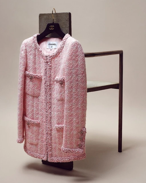 Pale pink Chanel jacket with four pockets and decorative trim, displayed on a hanger and chair against a neutral backdrop.