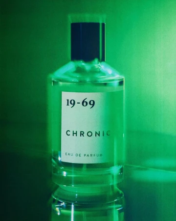 A clear glass perfume bottle with a dark cap and white label stands upright against a vibrant green gradient background.