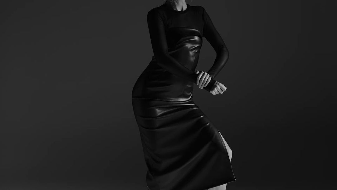 A woman poses in a studio wearing a long-sleeve black top and a black leather midi dress in a fashion portrait against a gray backdrop.