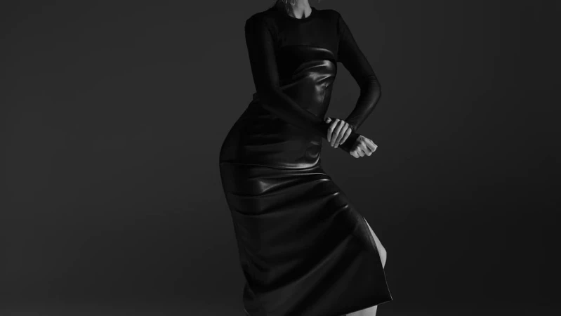 A woman poses in a studio wearing a long-sleeve black top and a black leather midi dress in a fashion portrait against a gray backdrop.