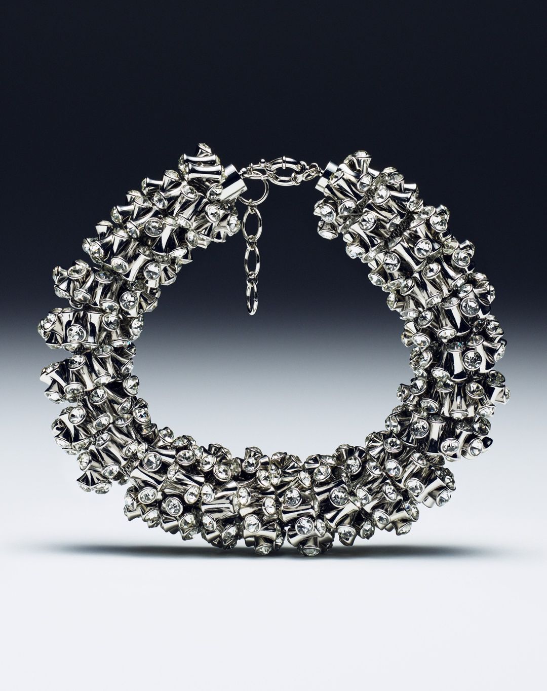 Close-up studio shot of a shiny silver choker necklace with diamond-like embellishments against a graduated gray backdrop.