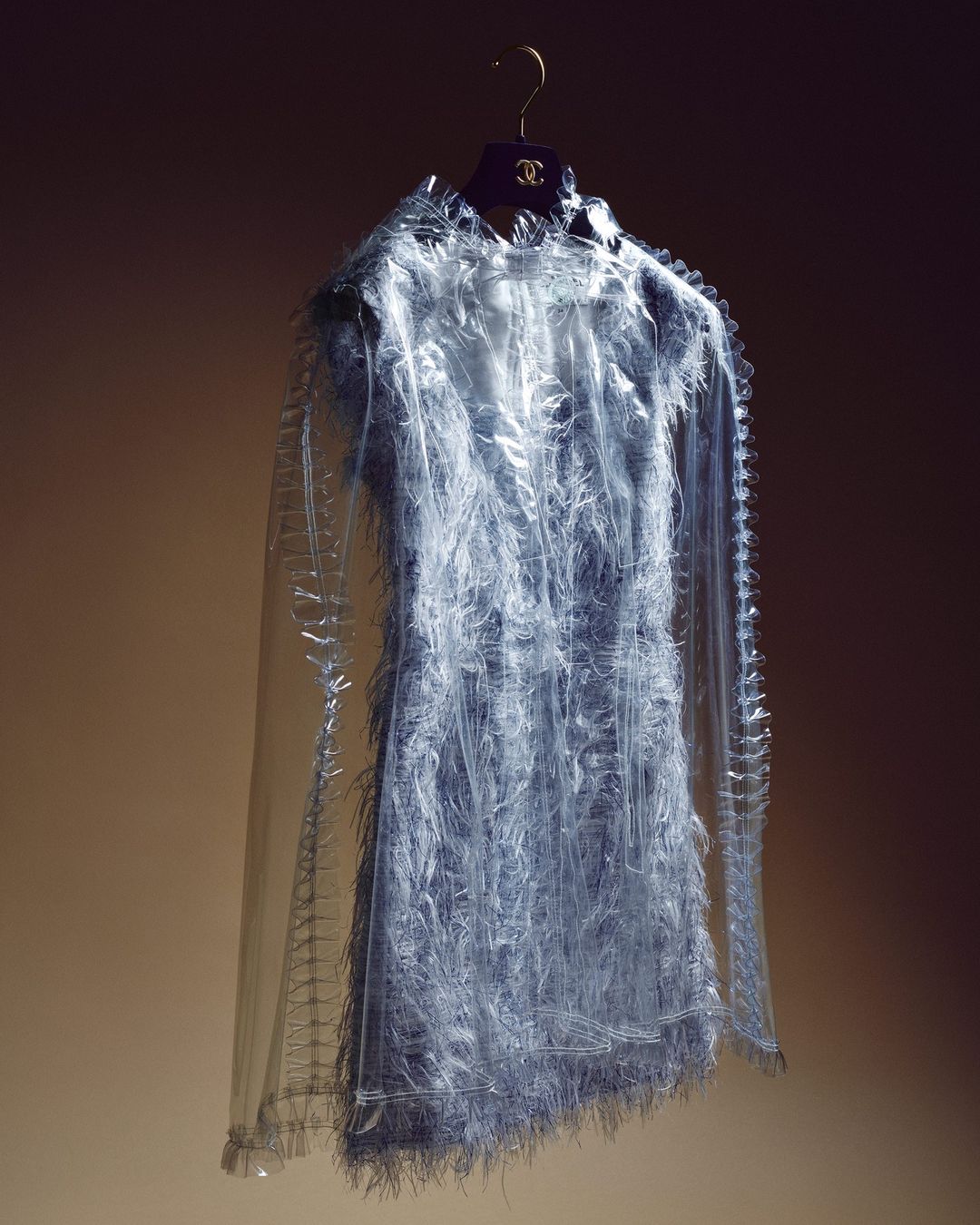 Fringe dress on hanger: a light blue, semi-sheer, fringed cocktail dress hangs against a graduated dark-to-light brown background.