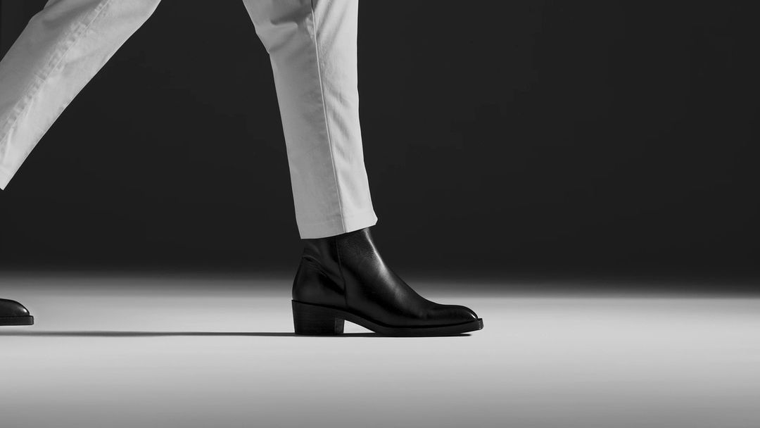 A person wearing white pants and black leather boots walks in front of a dark grey background with a light grey floor surface.