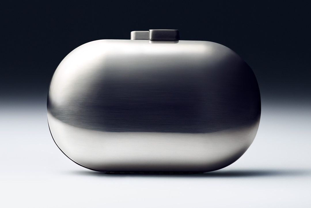Close-up shot of a futuristic silver clutch purse with rounded edges on a gradient gray background in a studio product photograph.