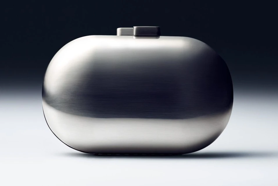 Close-up shot of a futuristic silver clutch purse with rounded edges on a gradient gray background in a studio product photograph.