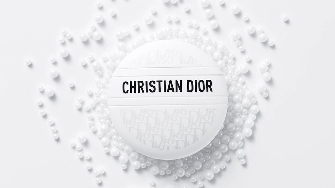 Close-up shot of white, round Christian Dior compact case surrounded by white beads against a white backdrop in a beauty product shot.
