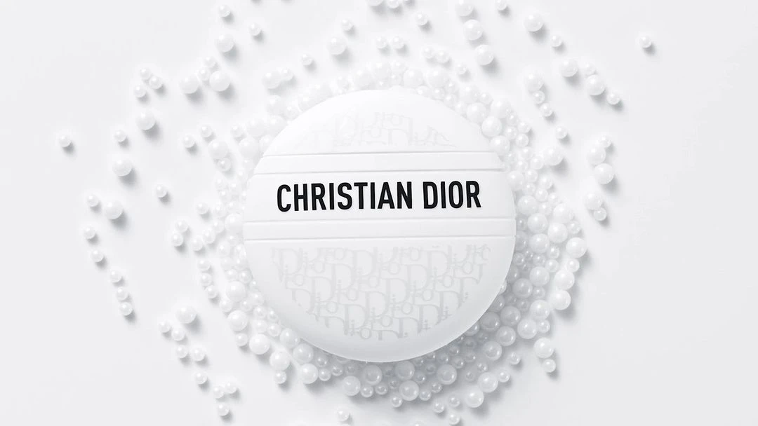 Close-up shot of white, round Christian Dior compact case surrounded by white beads against a white backdrop in a beauty product shot.