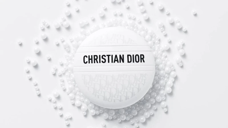 Close-up shot of white, round Christian Dior compact case surrounded by white beads against a white backdrop in a beauty product shot.