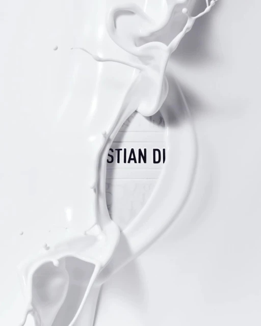 Dynamic splash of white liquid artfully surrounds a 'Christian Dior' sign, set against a bright white background, creating a minimalist aesthetic.