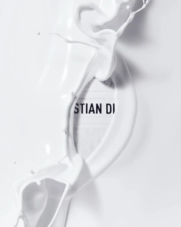 Dynamic splash of white liquid artfully surrounds a 'Christian Dior' sign, set against a bright white background, creating a minimalist aesthetic.