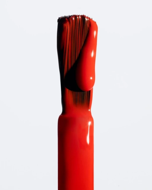 Macro studio shot of an applicator brush covered in bright red nail polish on a clean, white background with shadow detail.
