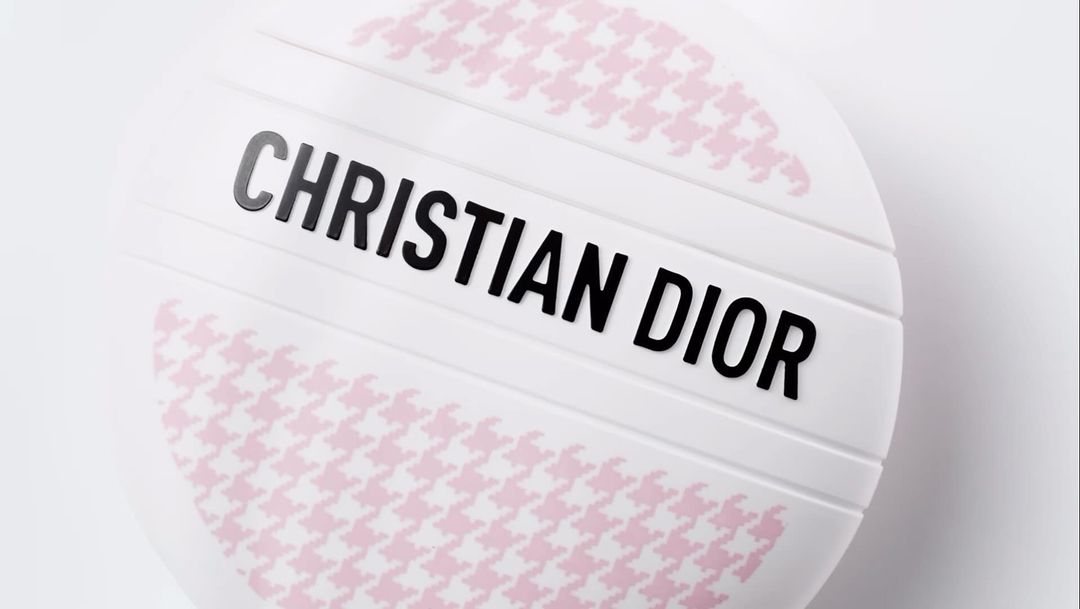 Close-up of a round, white makeup compact from Christian Dior with pink houndstooth pattern against a white background.