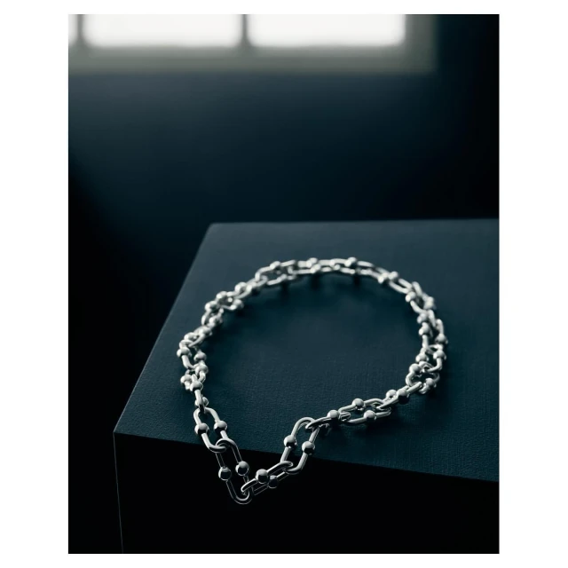 Silver chain-link necklace on a black surface, softly lit from the side, set against a dark background with a blurred window.