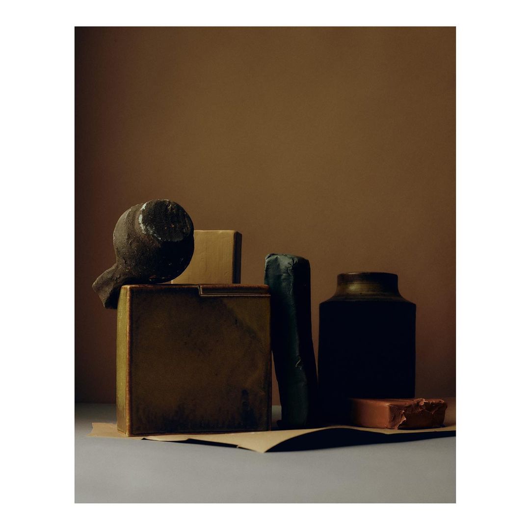 Still life composition features dark pottery jug, soap bars, small block, and brown paper on a grey surface against a brown backdrop.