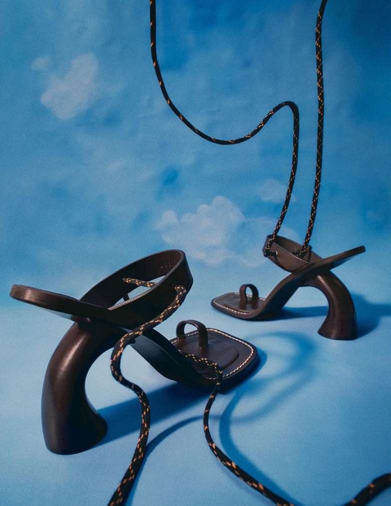 Two black high-heel sandals with toe loops and braided ankle straps are showcased against a soft blue background with cloud patterns.