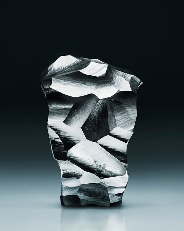 Abstract sculpture with crystalline facets and gray textured surface, contrasted against a dark gray to light gradient background.