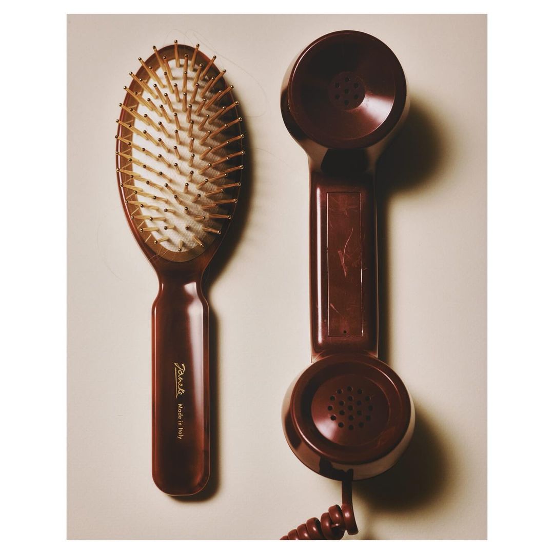 Still life photograph of a brown hairbrush and a vintage brown telephone receiver against a neutral background, lit by soft light.