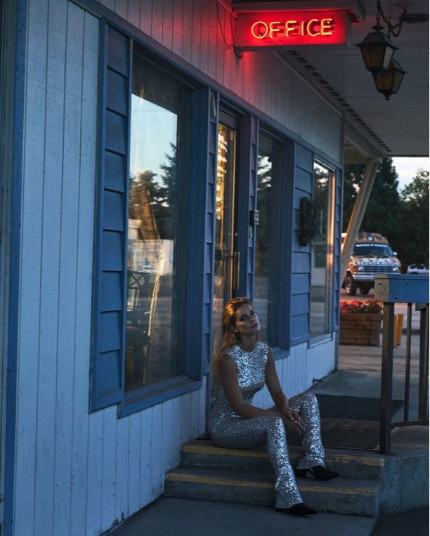 Woman in a sparkly outfit sits on the steps of a blue building with an 'office' neon sign at dusk. A vintage car is parked nearby.