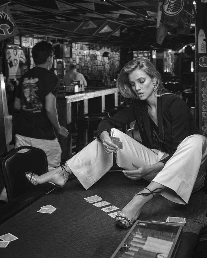 Monochrome shot of a woman sitting cross-legged on a table in a bar holding playing cards, with another man standing in the background.