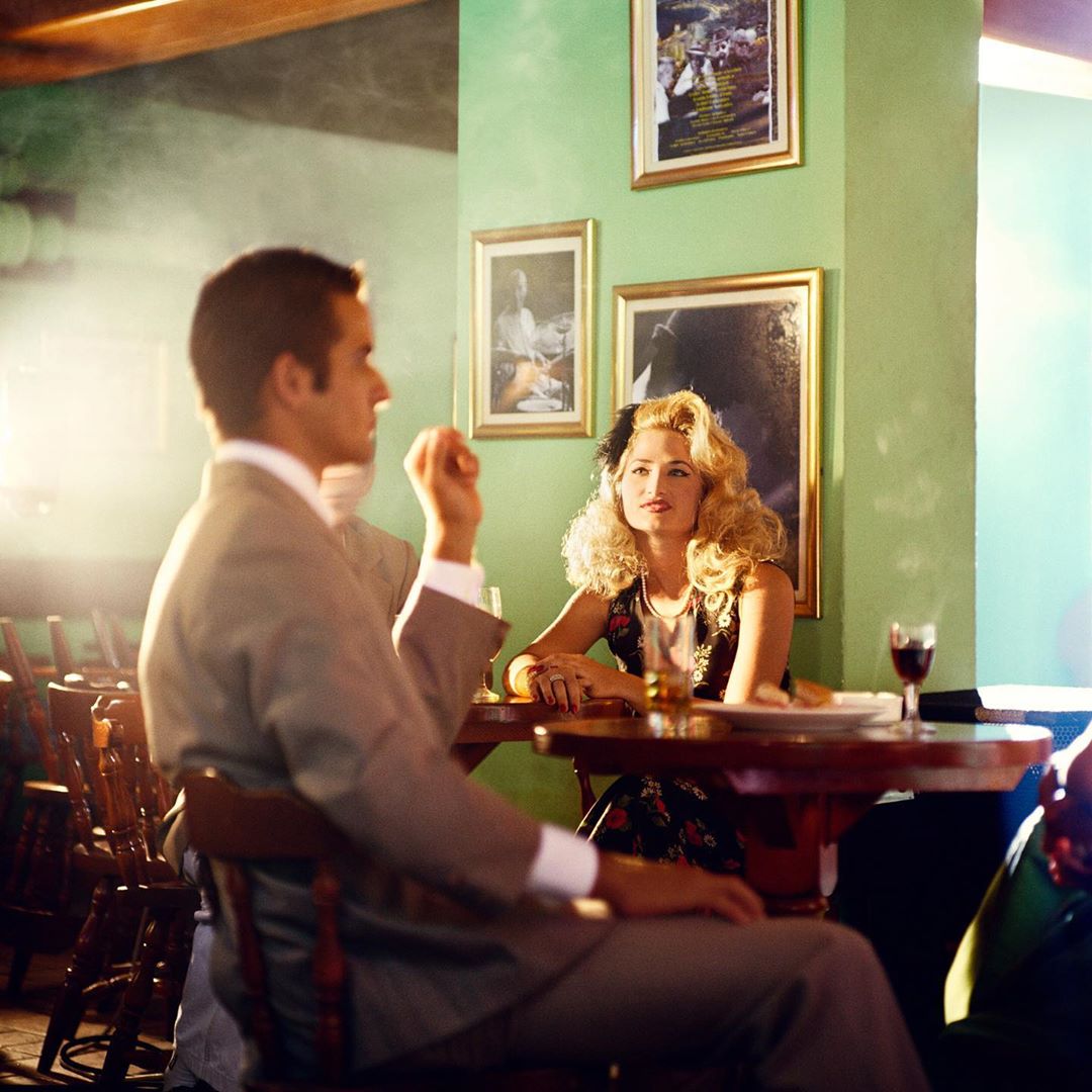 Woman and man at a table in a bar or restaurant with paintings, glasses, and plates; soft lighting and vintage style.