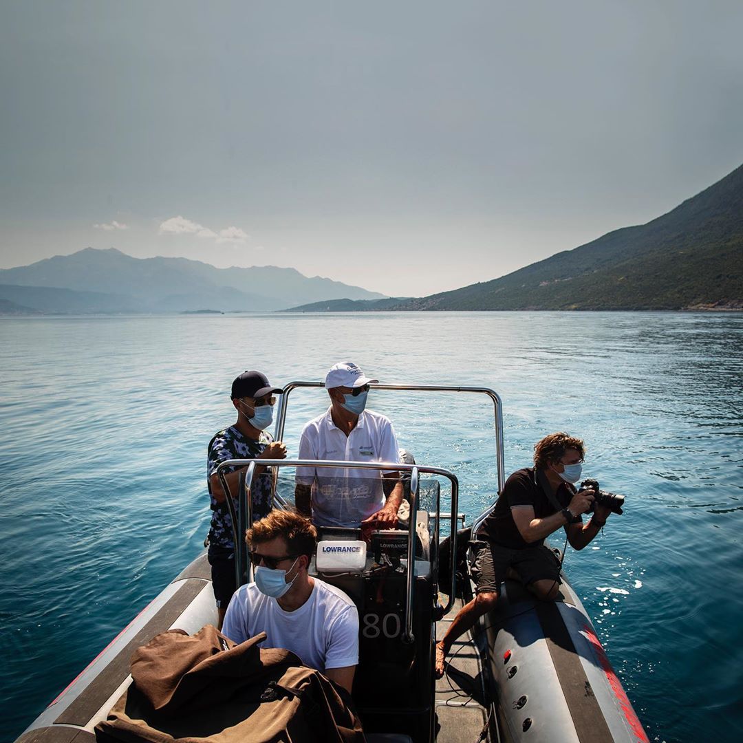 Four people, all wearing face masks, ride in an inflatable boat on the ocean near a mountainous coastline on a clear sunny day.