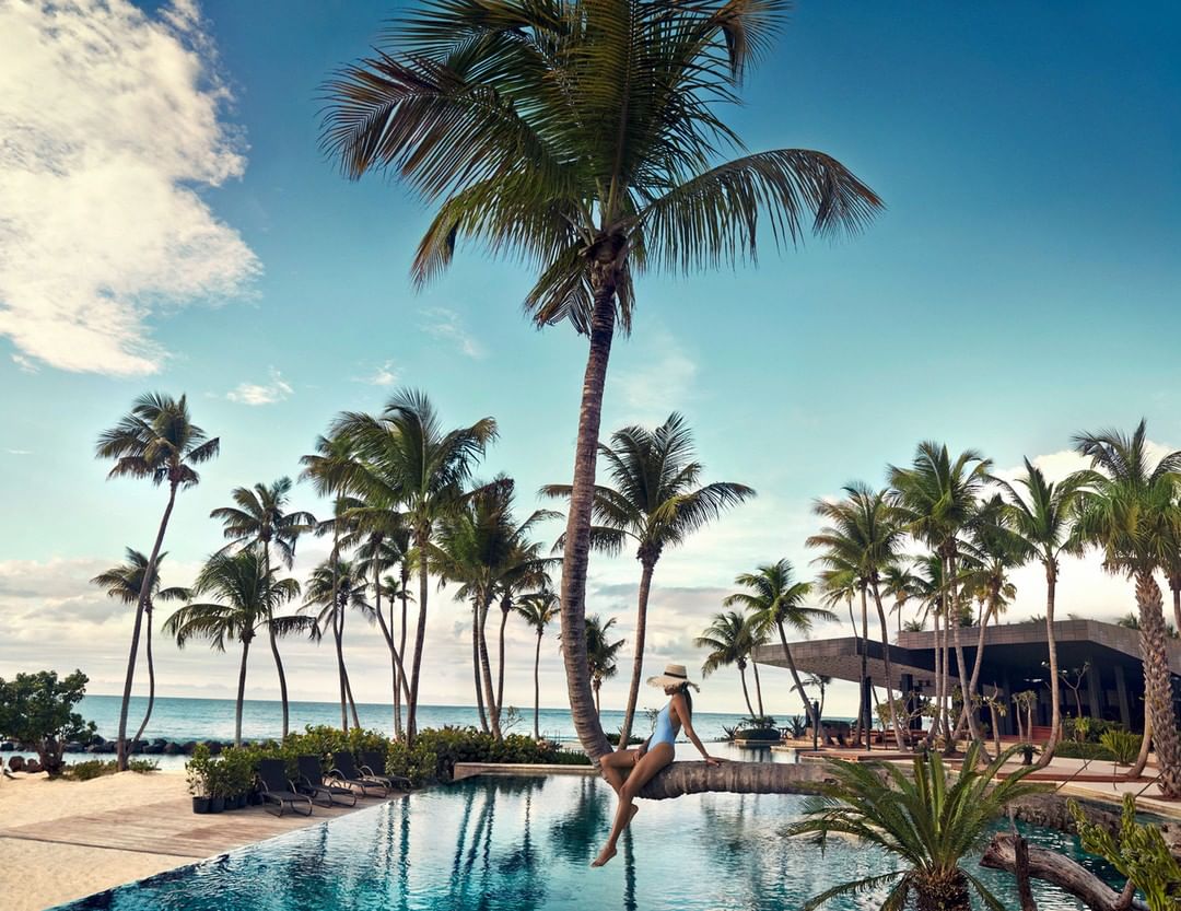 Woman in swimwear and sunhat sits on a palm tree trunk overlooking an infinity pool and tropical beach with palm trees against a blue sky.