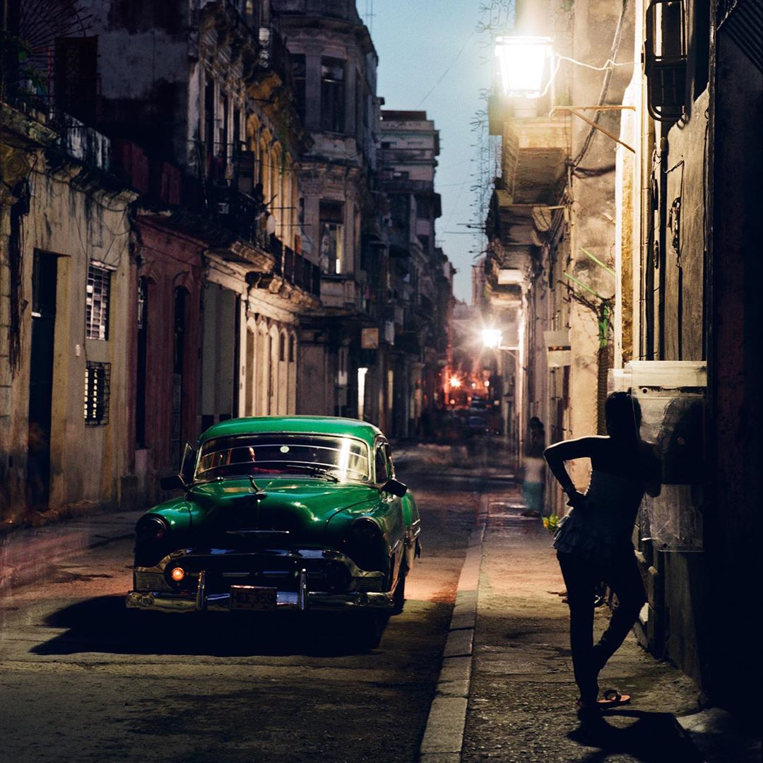 Vintage green car parked on a Havana, Cuba street, with a woman standing nearby, under dim streetlights against an old building backdrop.