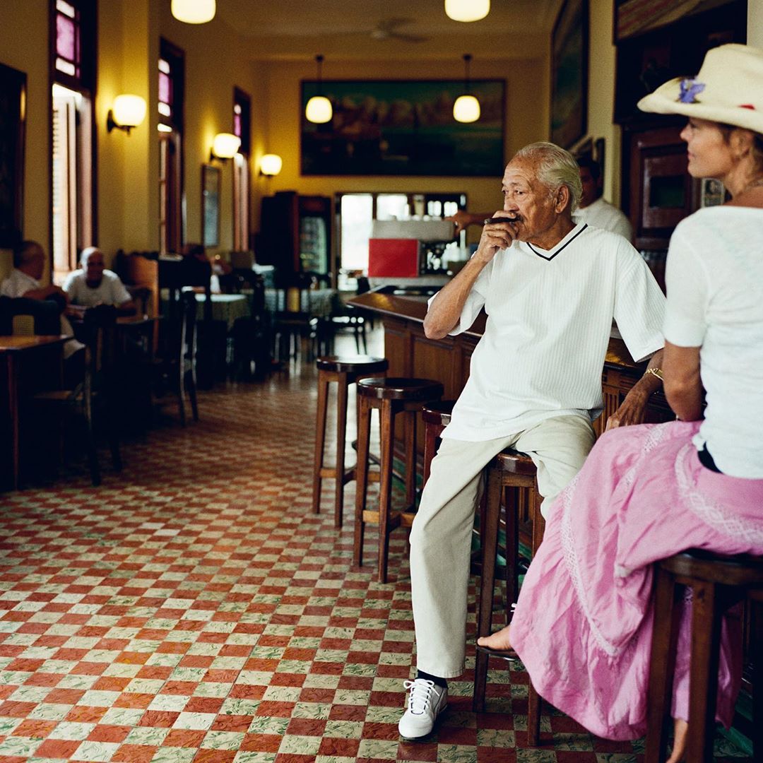 Two people sitting at a bar inside an old-fashioned interior with red and white checkered flooring, dark wood bar, and overhead lighting.