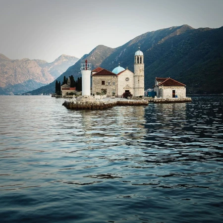 Island with a church and lighthouse in the middle of the water, surrounded by mountains and clear skies, creating a serene landscape.