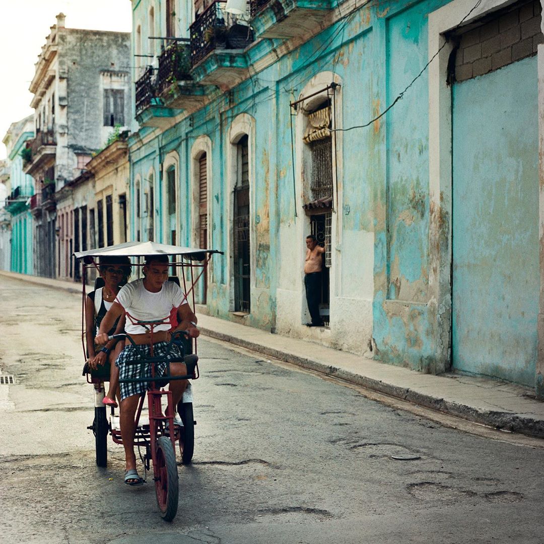 A red pedicab with two people travels down a narrow city street lined with buildings in a somewhat run-down urban setting.