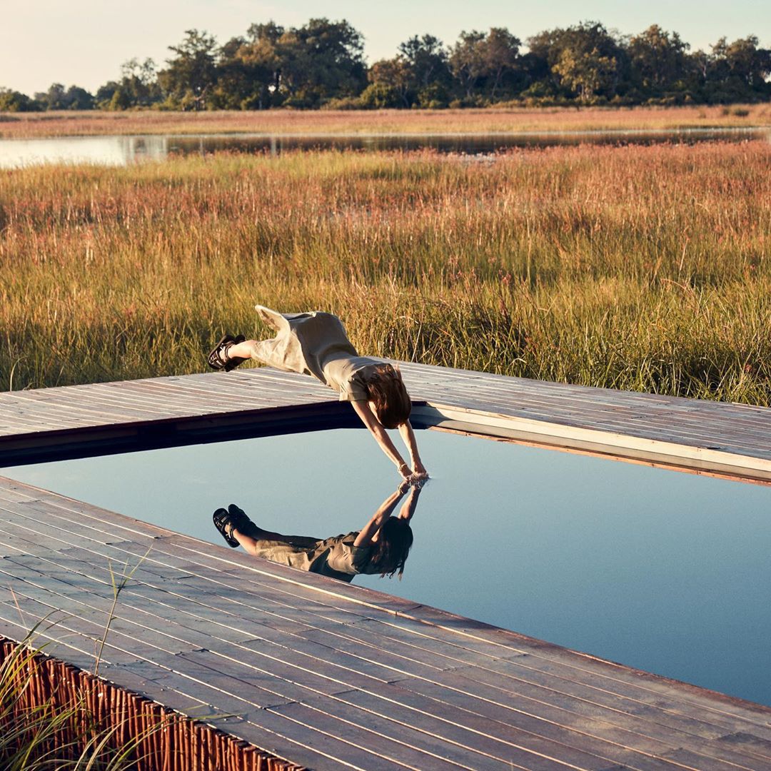 Woman dips into pool with tall grass and trees in background, reflecting in pool water, showcasing outdoor luxury travel and fitness.