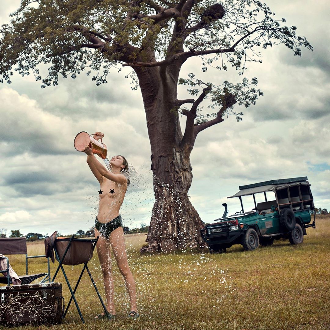 Adult female outdoors pouring water on herself from a copper jug, wearing a bikini bottom, with a safari car parked in the background.