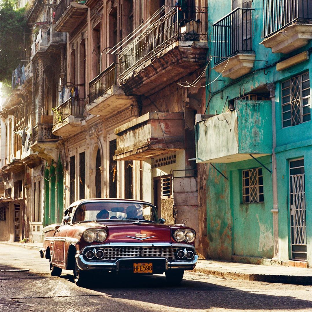Maroon vintage car parked on a sunny street in Havana, Cuba, with colorful buildings and a classic urban scene.