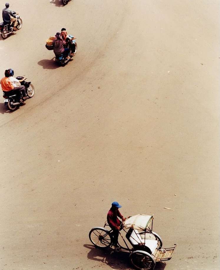 Overhead view of a man pedaling a cyclo on a tan road, surrounded by scattered motorcycles on a sunny day. Transport and urban scene.