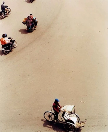 Overhead view of a man pedaling a cyclo on a tan road, surrounded by scattered motorcycles on a sunny day. Transport and urban scene.