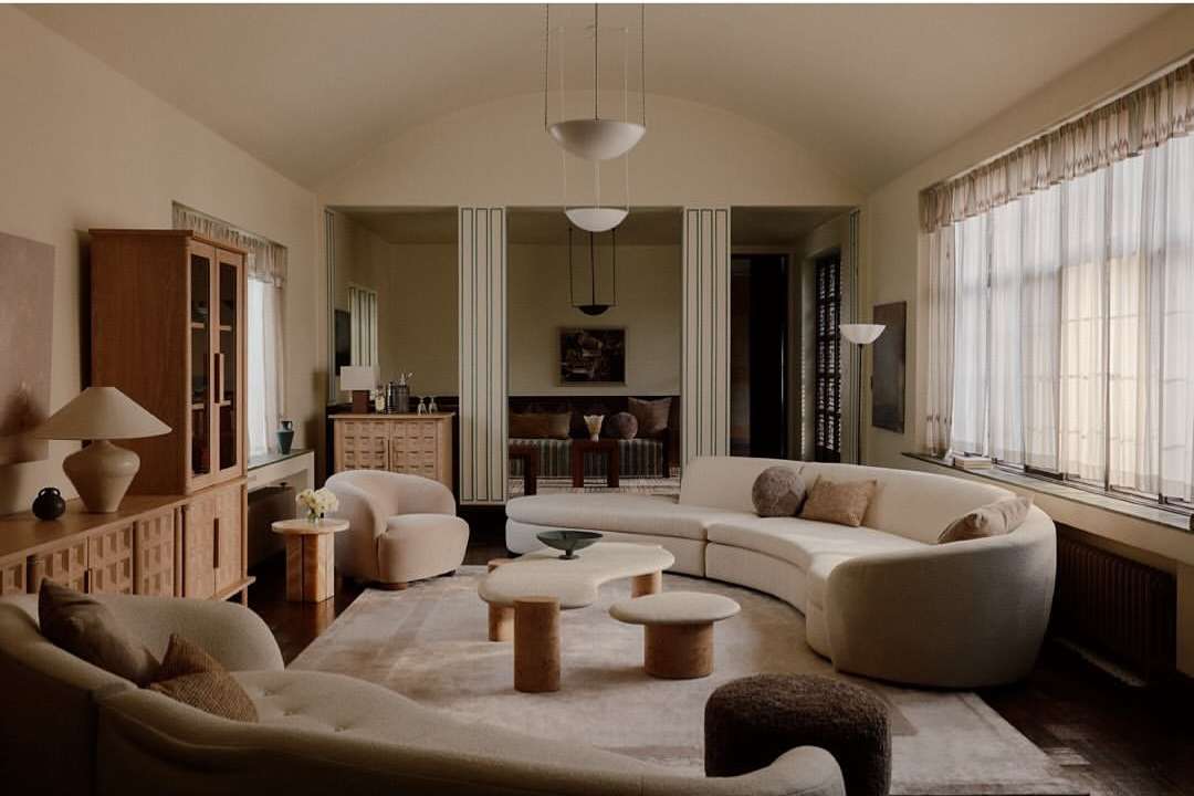 Living room with curved sofas, wooden cabinets, mushroom tables, soft rug, overhead lighting and neutral color palette in home interior.