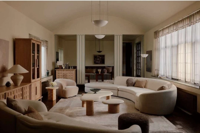 Living room with curved sofas, wooden cabinets, mushroom tables, soft rug, overhead lighting and neutral color palette in home interior.