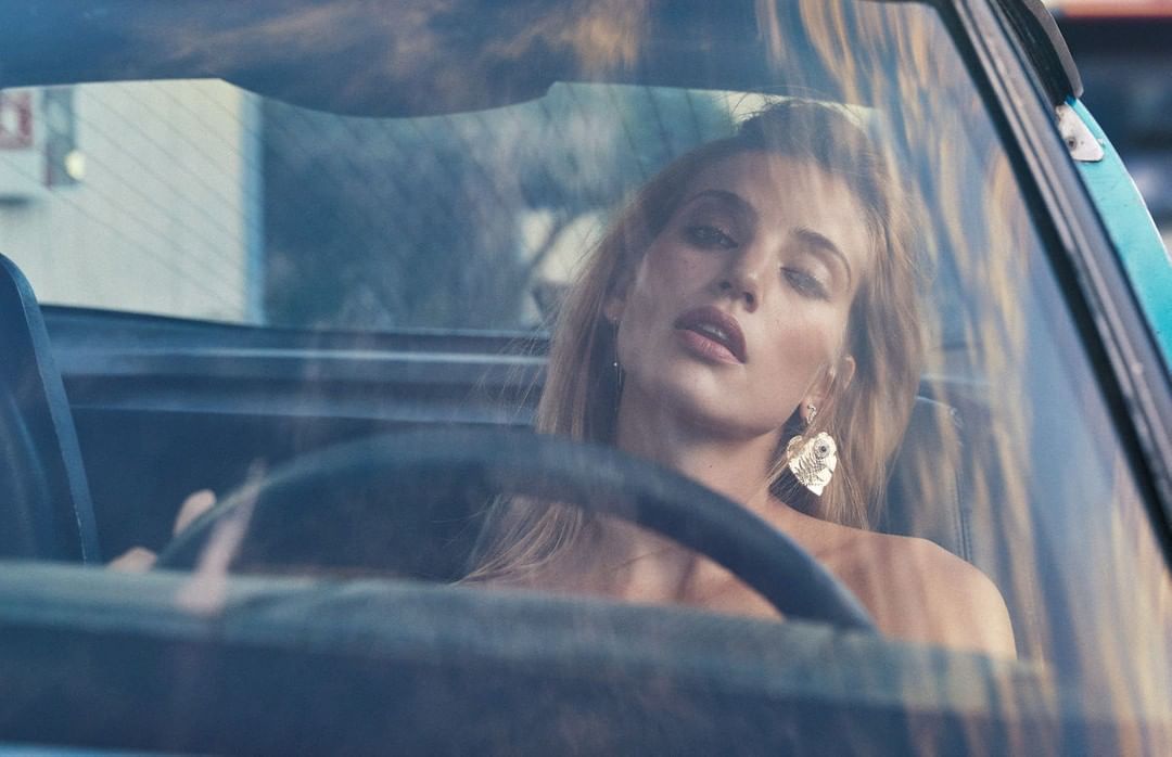 Blonde adult female poses inside a vintage car with a blue exterior and an outdoor background scene, golden jewelry and hair.