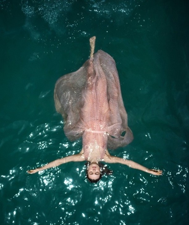 Woman in an elegant pink dress floats on her back in teal water, creating a serene and surreal image with a dreamlike quality.